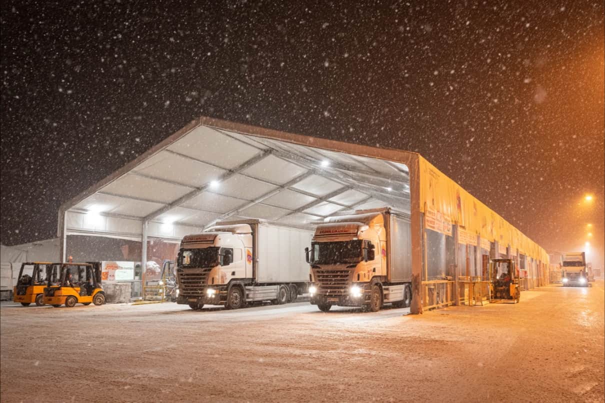 Kar yağışı altında gece, tırların yanaştığı aydınlatmalı lojistik çadırı