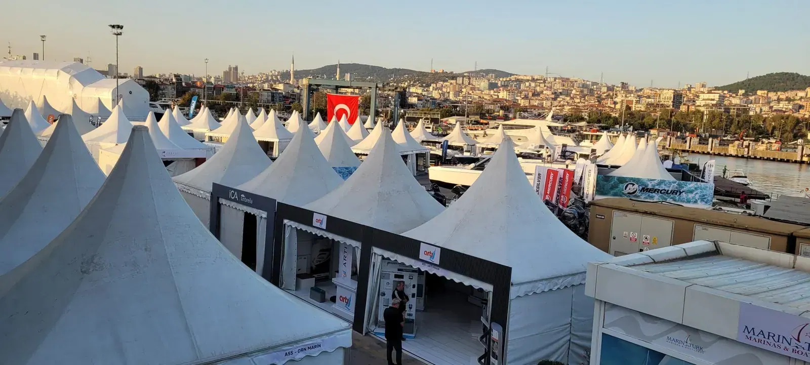 Marina kıyısında sıralanmış beyaz pagoda çadırları ve arkada şehir manzarası