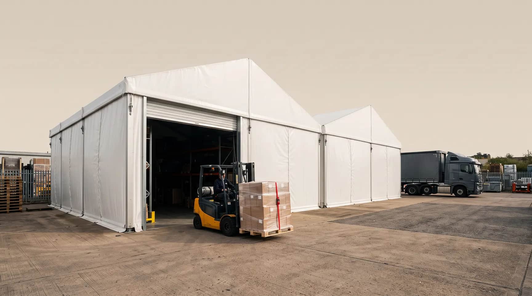 Sıra halinde iki beyaz depo çadırı, açık rulo kapıdan paletle çıkan forklift, sabah ışığı