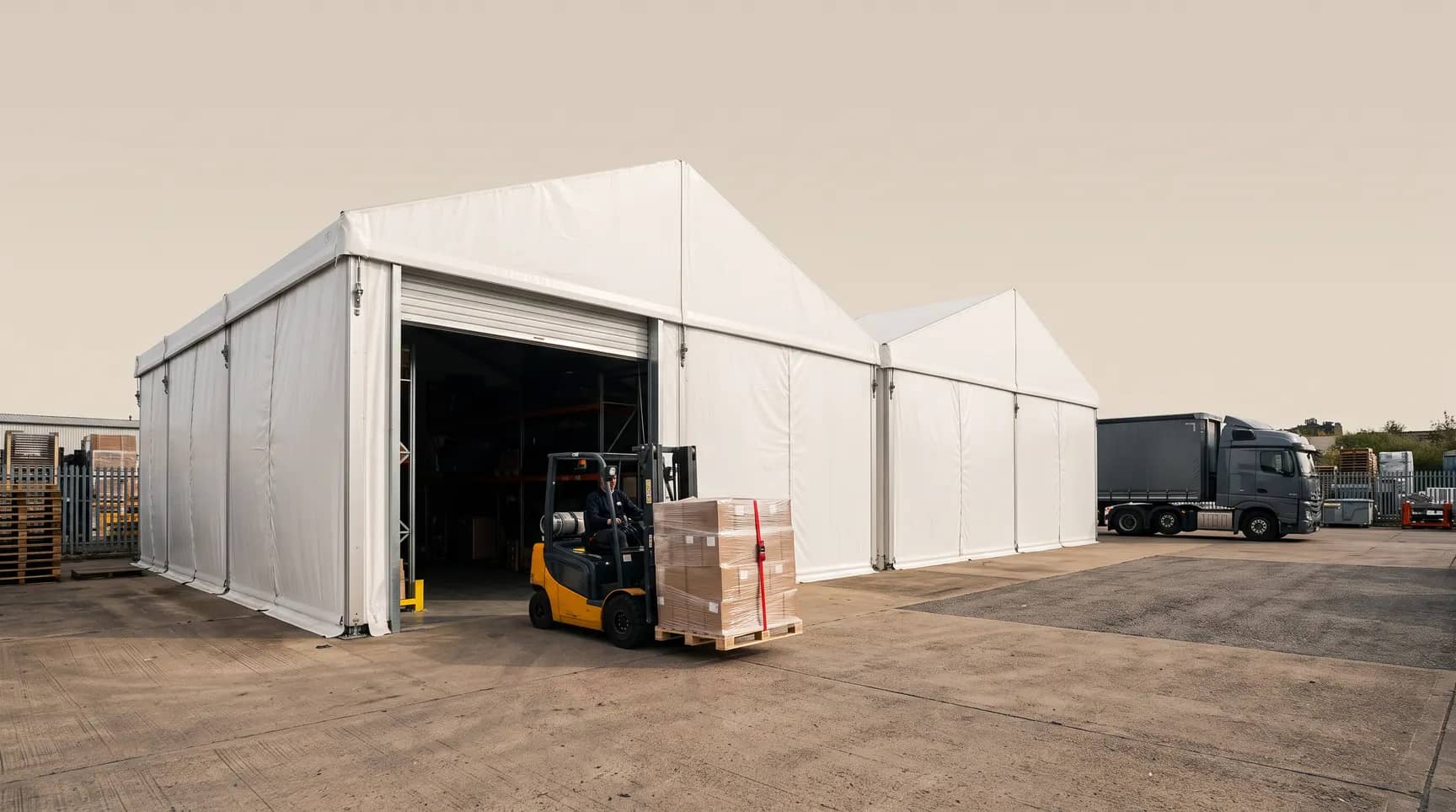 Sıra halinde iki beyaz depo çadırı, açık rulo kapıdan paletle çıkan forklift, sabah ışığı