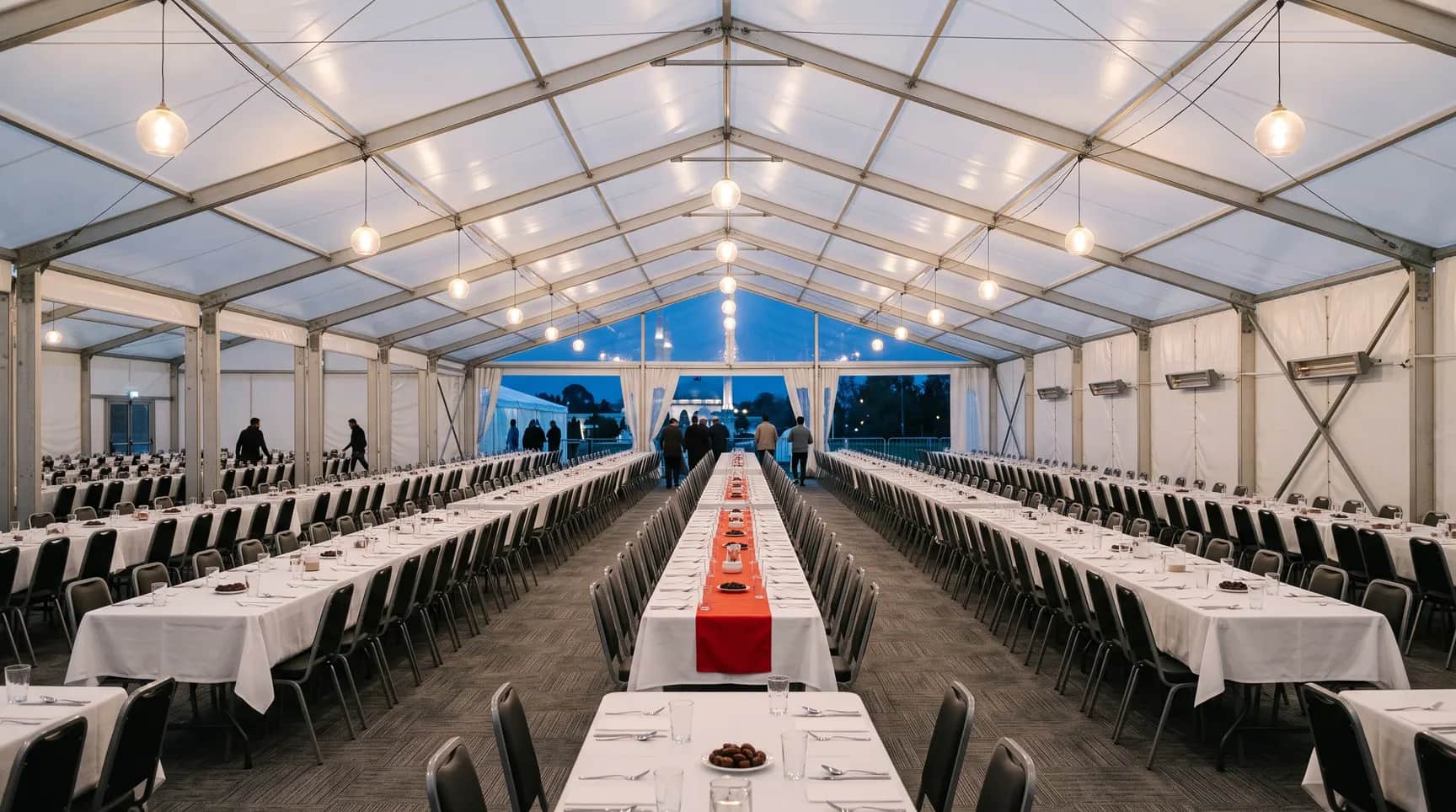 Büyük iftar çadırı iç mekânı: uzun masa sıraları, sıcak aydınlatma ve dışarıda mavi saat
